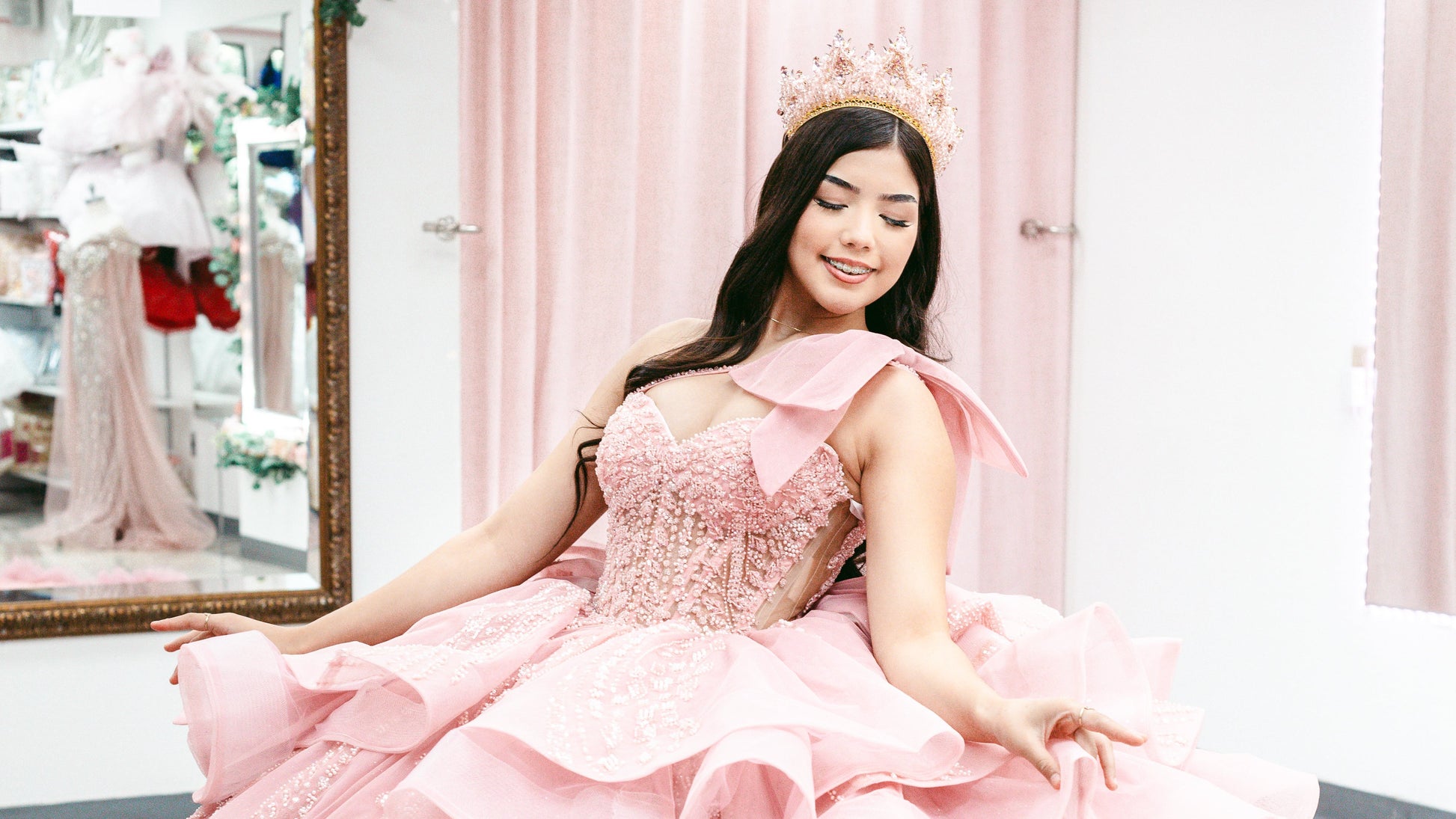 A woman wearing a voluminous light pink ball gown with layered ruffles, sequined details, and a matching pink crown, standing in a boutique with pink curtains and a mirror in the background.