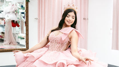 A woman wearing a voluminous light pink ball gown with layered ruffles, sequined details, and a matching pink crown, standing in a boutique with pink curtains and a mirror in the background.