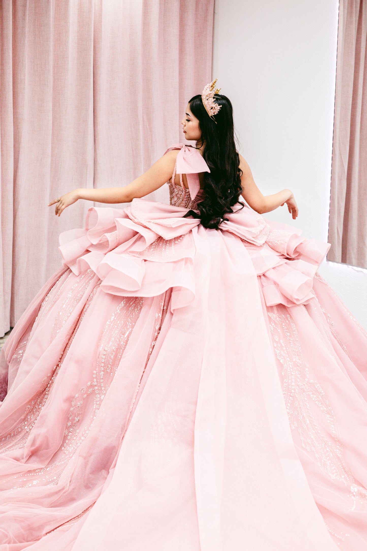 A woman wearing a voluminous pink ball gown with a large ruffled bow at the back, posed indoors against pink curtains and a white wall.